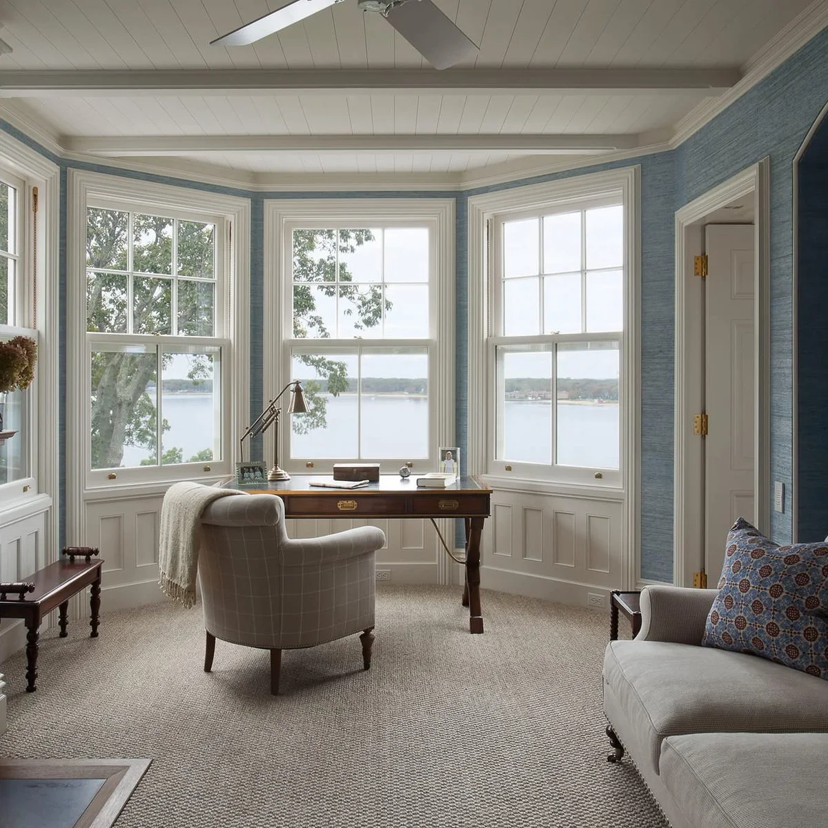 A wooden desk and upholstered chair within a blue-walled bay with three windows overlooking water.
