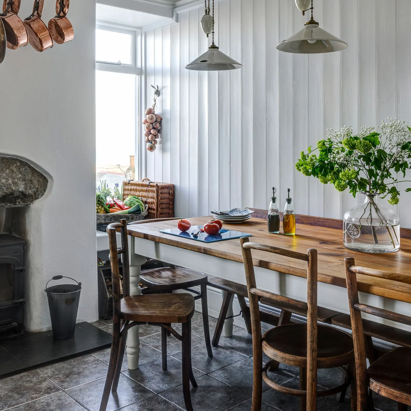 Cottage dining corner with pale vertical wall paneling, a wooden table, darker wooden chairs, two pale pendant lights and a narrow window.