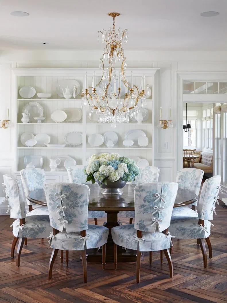Large white fitted shelves displaying pale dishes above closed lower cupboards, with a round dark table, blue-patterned chairs, flowers, and an ornate chandelier.