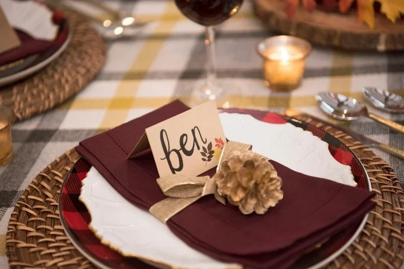 A handwritten name card and small pinecone sit over a plum napkin on a pale plate above a woven charger and checked cloth