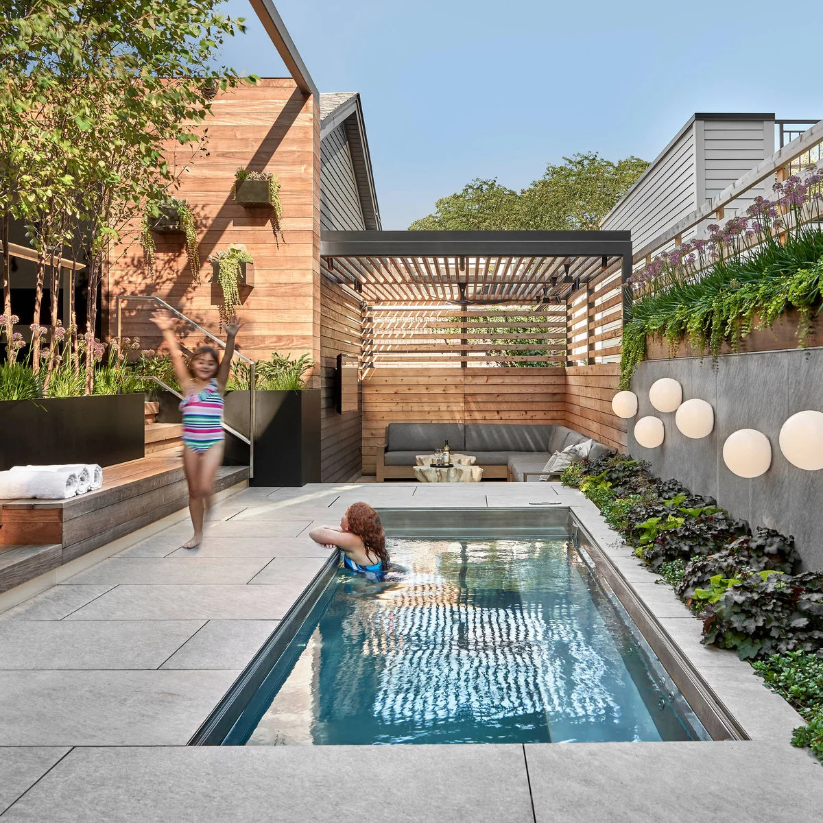 A narrow rectangular pool runs beside gray paving, raised planting and wood fences, with covered seating at the far end.