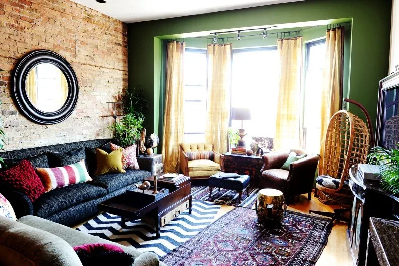 Green living room with a brick wall, dark sofa, hanging chair and layered patterned rugs.