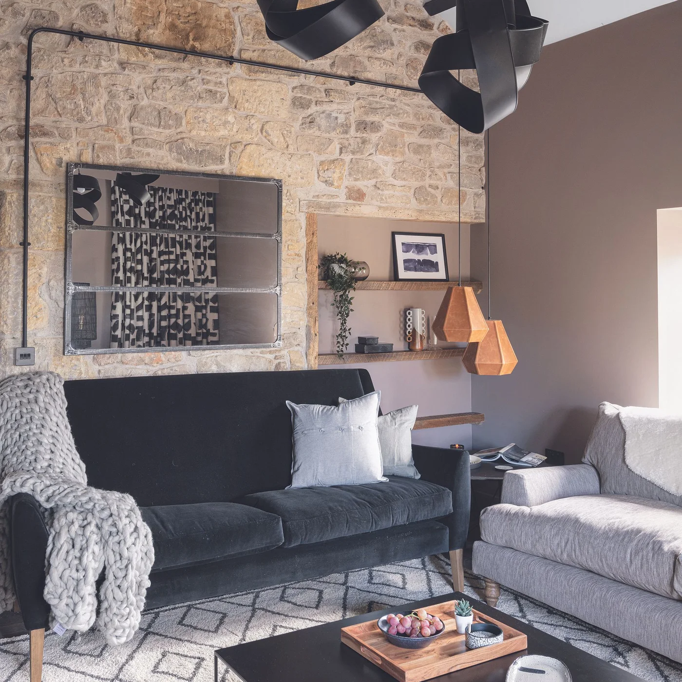 A pale rough masonry wall with framed art stands behind a dark sofa beside wooden recessed shelves and a gray armchair