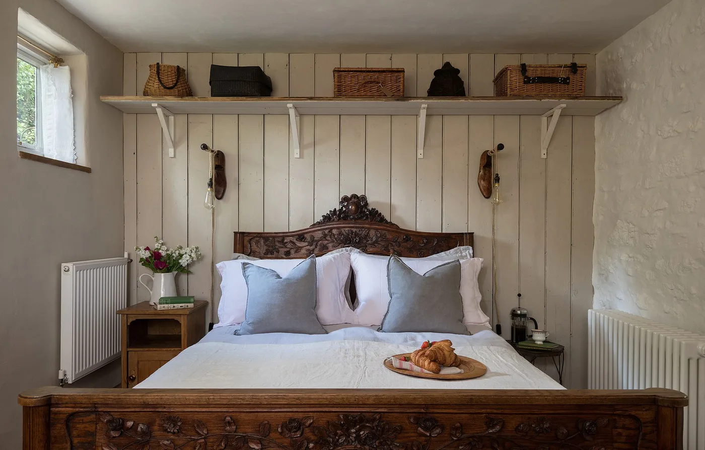 A carved timber bed stands against pale vertical boards beneath a long shelf holding woven bags and baskets.
