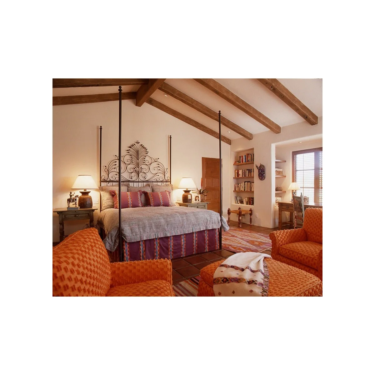 Tall metal bed with a striped bed skirt and pillows, orange chairs and a woven ottoman beneath exposed wood beams.