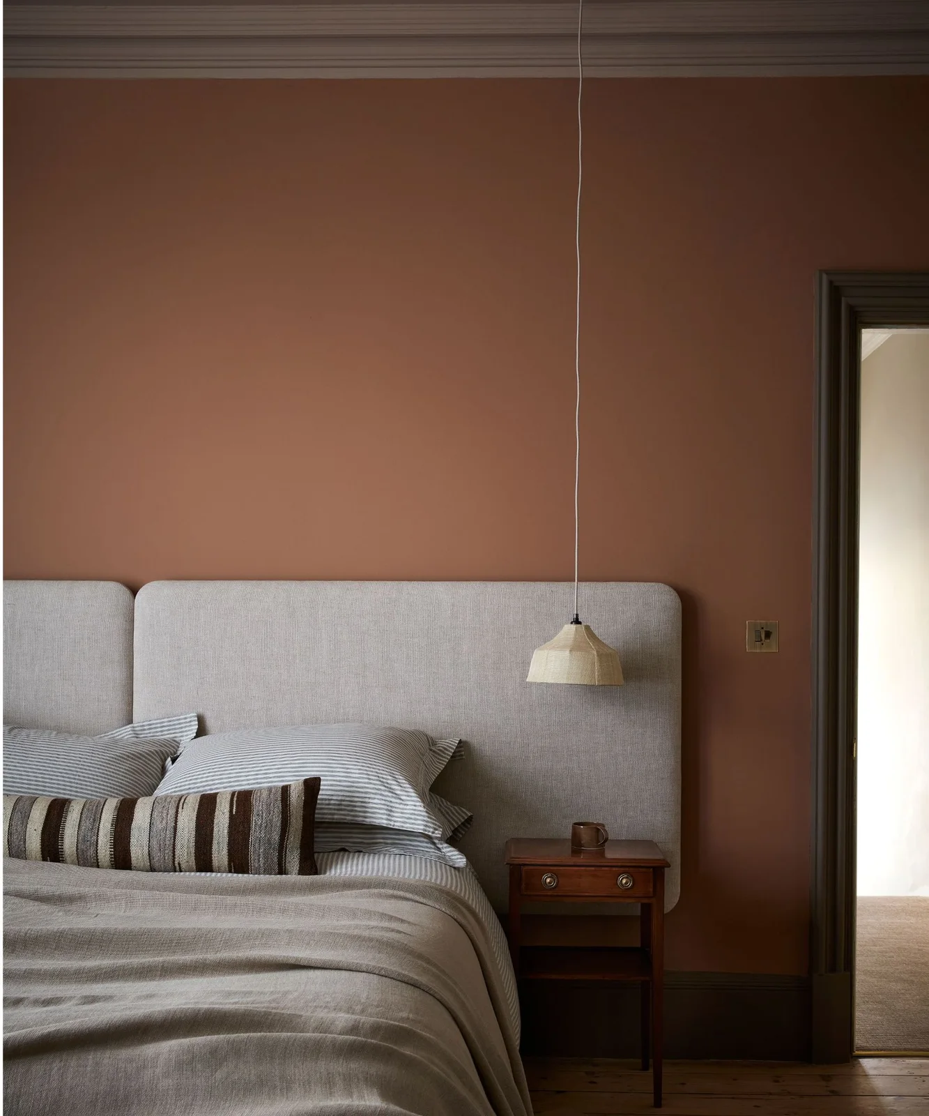 A pale upholstered headboard stands against a warm pink wall beside striped bedding, a wooden drawer cabinet and a small hanging light