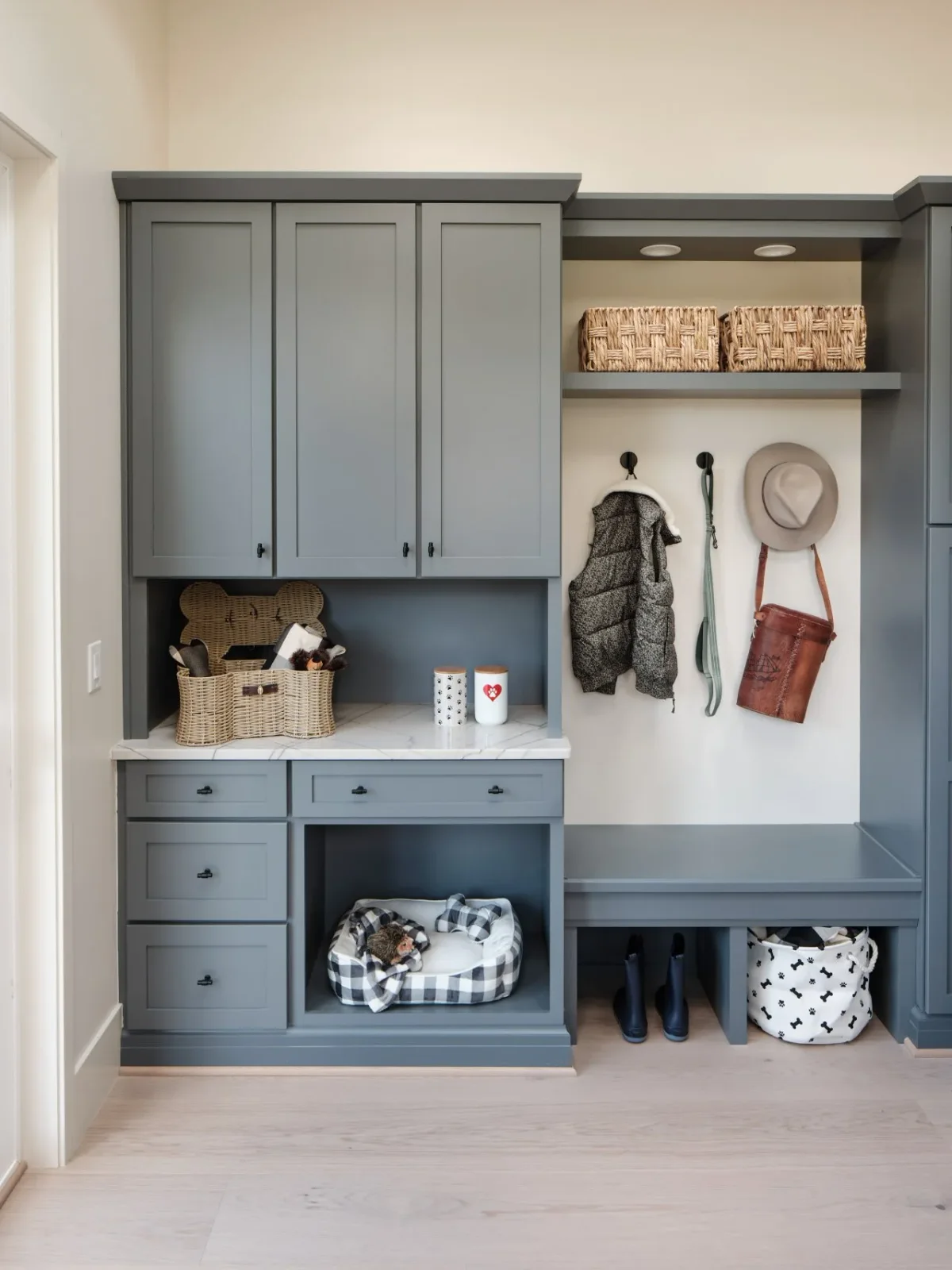 Gray mudroom cabinetry includes upper cupboards, small drawers, a pet-bed niche and an open bench with coat hooks and baskets.