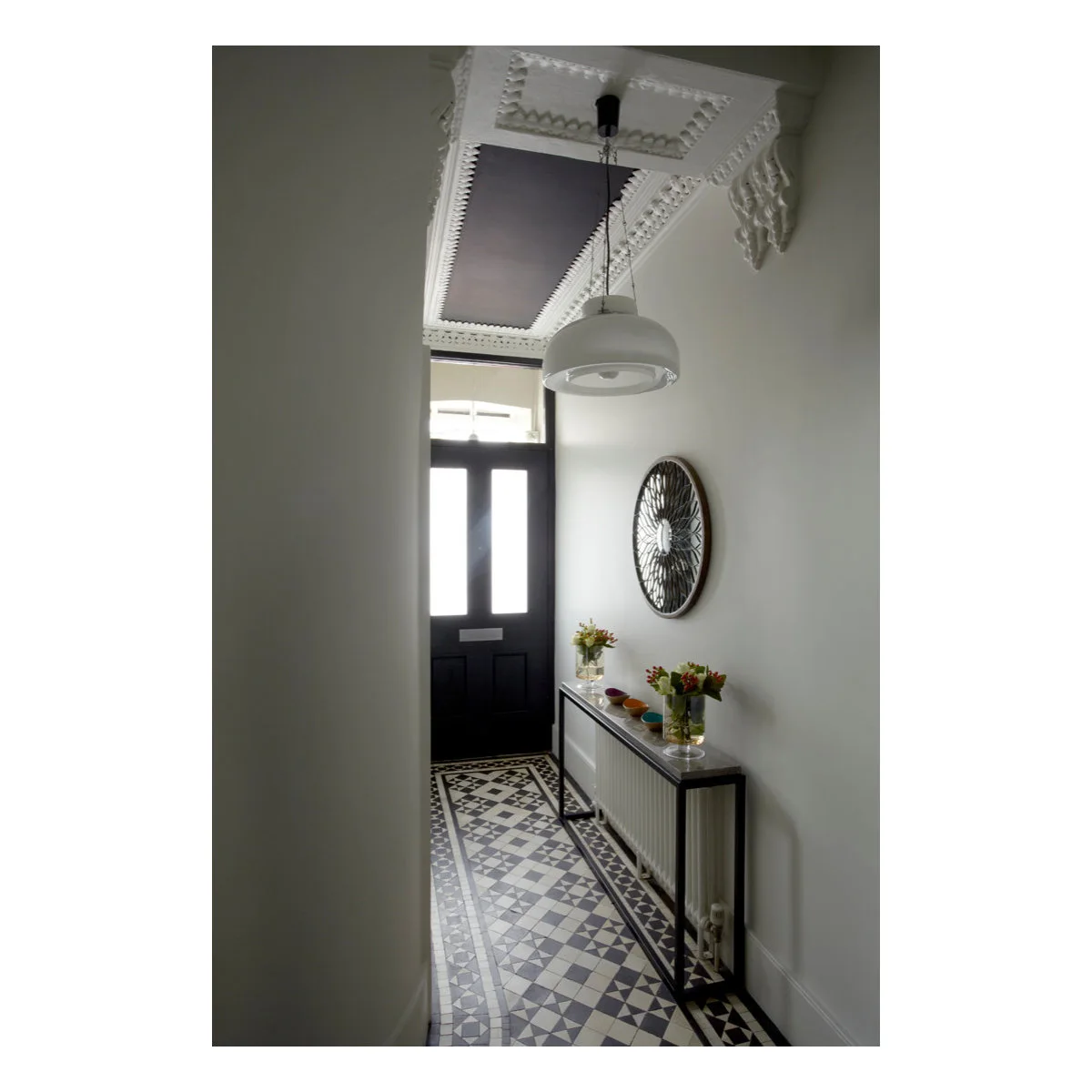 Narrow entrance with a black door, patterned floor, round mirror and a slim console positioned over a radiator.