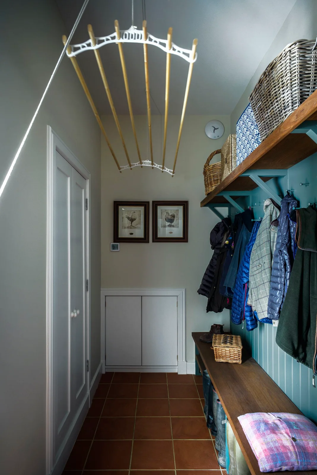 A narrow boot room has a blue paneled bench wall with hanging coats, high baskets and a suspended drying rack above a tiled central passage.