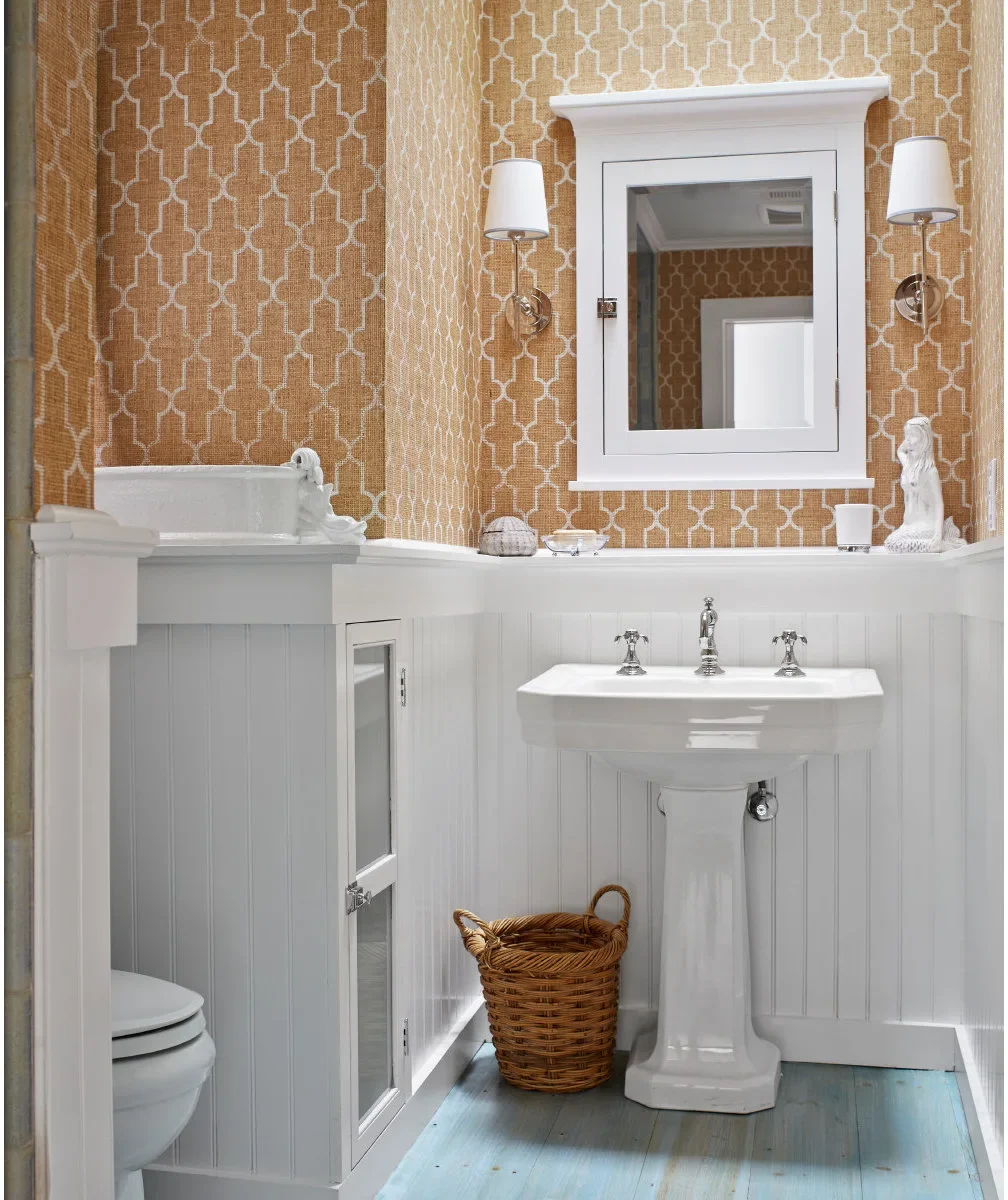 A white pedestal sink stands beside a narrow built-in cupboard below orange patterned walls, with pale blue painted floorboards.