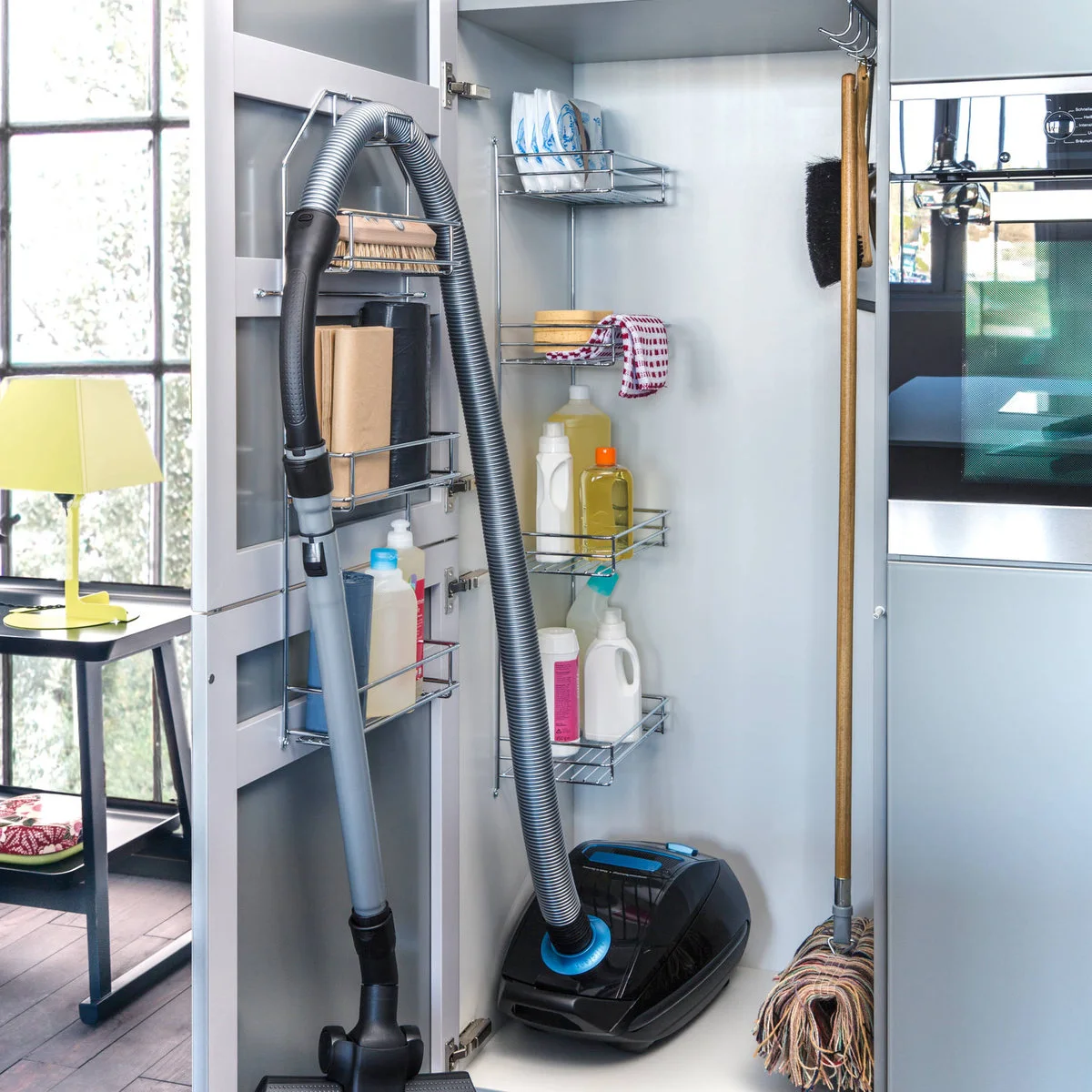 Tall kitchen utility cupboard with vacuum hose, mop and wire racks of cleaning products