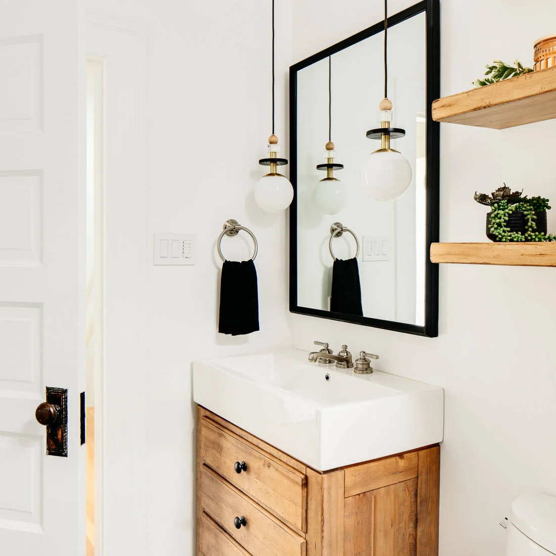 A small wood drawer vanity supports a white deep-sided basin below a black-framed mirror, globe pendant lights and simple wood shelves.