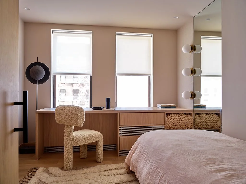A long wood desk beneath two windows stands beside a pale pink bed and textured light chair with mirror panels and small globe lights nearby