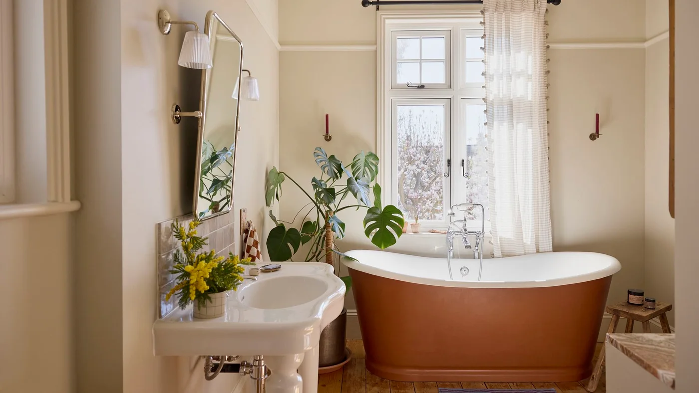 Warm terracotta-colored freestanding bath beside a white basin, pale walls, a long curtain and a small wood stool.