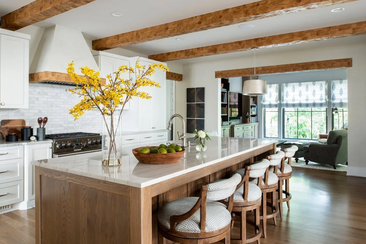 A natural oak island with pale counters and four upholstered stools sits before white kitchen cupboards beneath exposed wood beams.