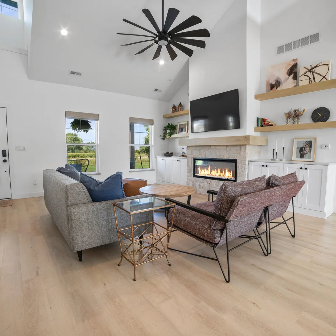 A pale room has a long horizontal fireplace with a light textured surround, wood mantel and shelves, white lower cupboards, gray seating and a small round wood table.