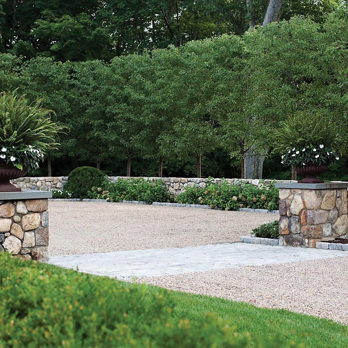 Gravel entrance between two stone piers topped with planted urns, with a low wall and clipped green planting beyond.