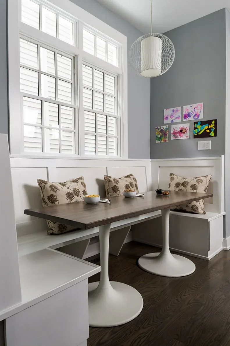 White paneled corner banquette beneath shuttered windows, with patterned pillows, a dark rectangular tabletop on two white pedestal bases, gray upper walls, and children’s art.