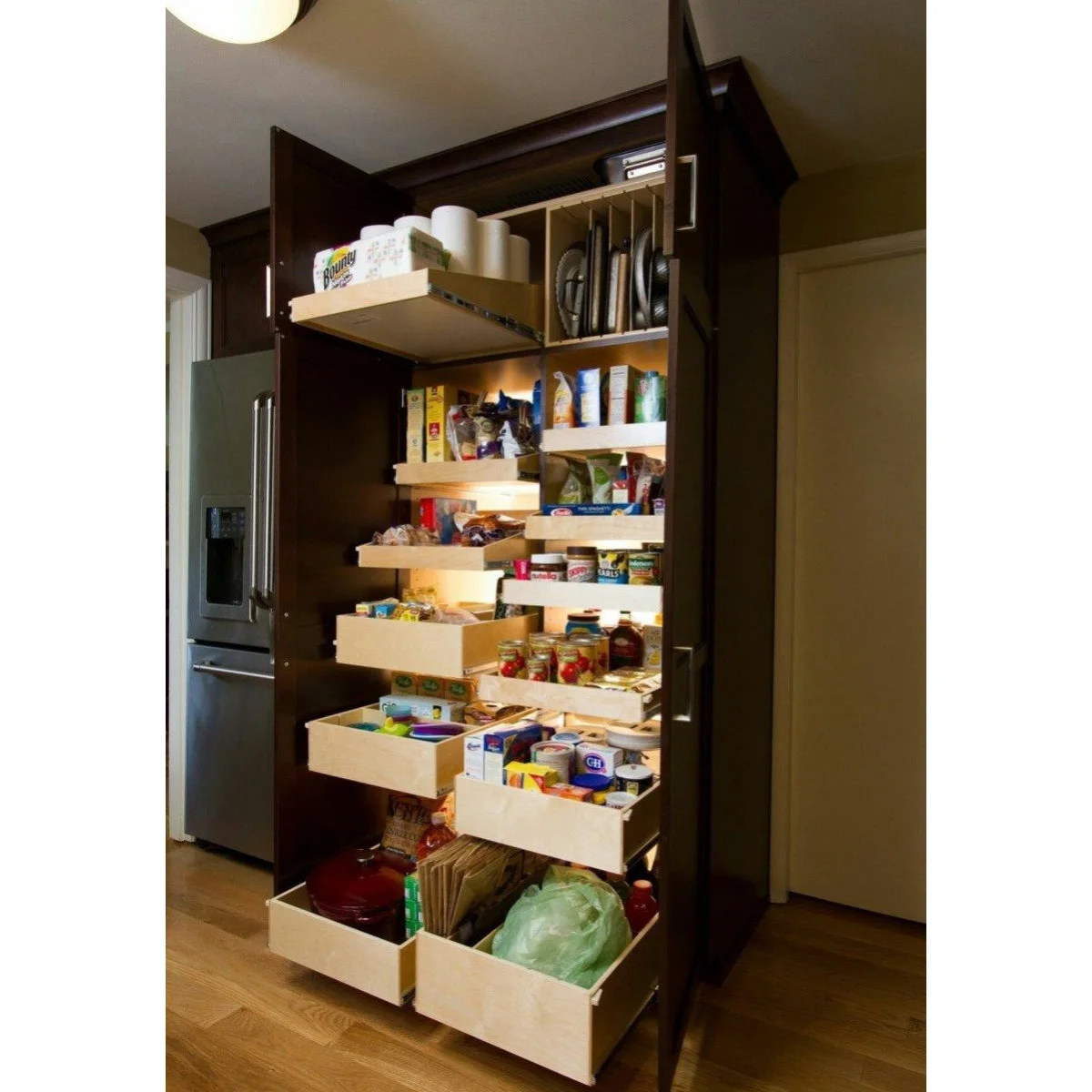 A tall dark wood cupboard opens beside a refrigerator to show pull-out trays, lower drawers and upright slots for trays or boards.
