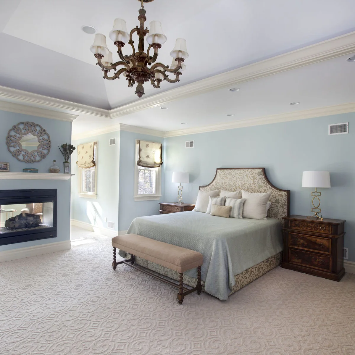 A pale blue bedroom with a patterned headboard, wooden bedside chests, shaded lamps and an upholstered bench.