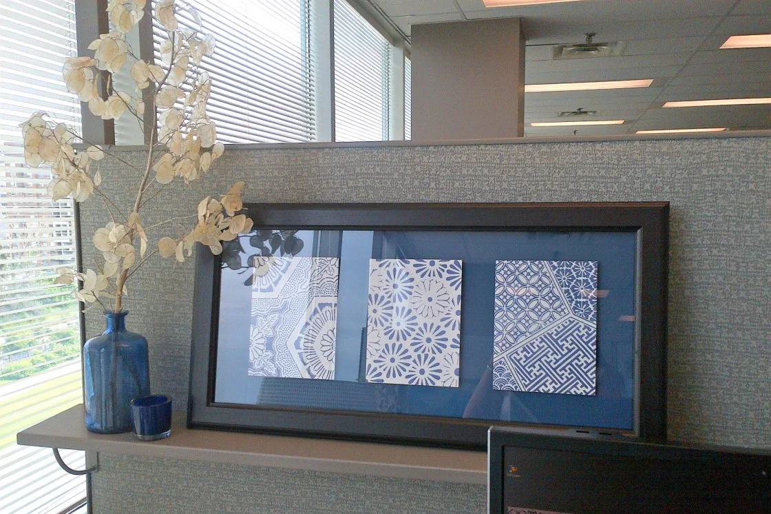 Wide dark frame containing three blue and white patterned papers on a shelf against a gray cubicle panel beside a blue vase.