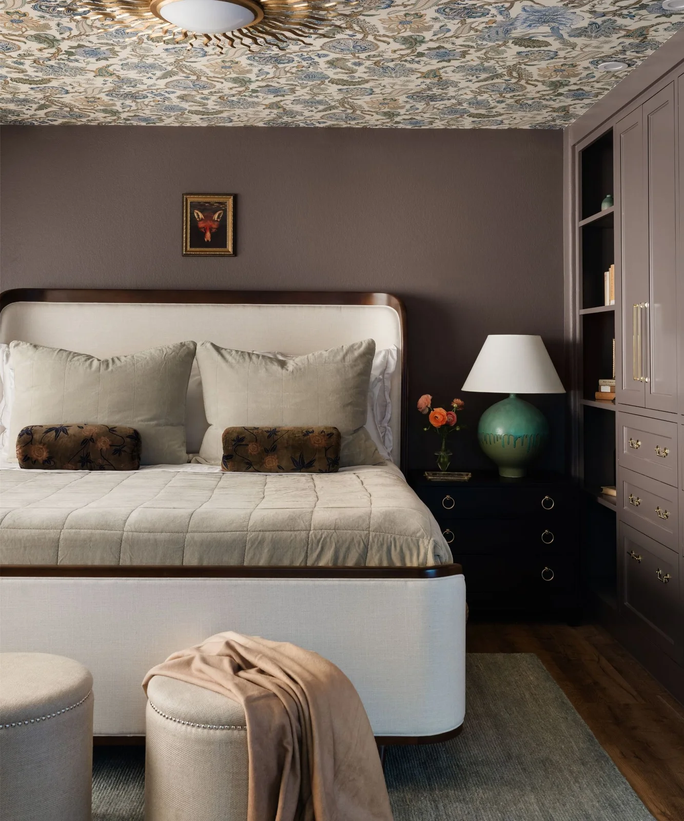 Mushroom-colored bedroom wall and matching fitted cupboards beside a pale upholstered bed and green lamp.