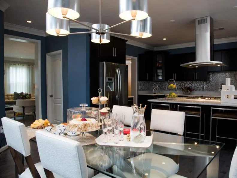 Rectangular clear glass dining table with pale chairs and a tray of glasses in a blue-walled kitchen with black cabinets
