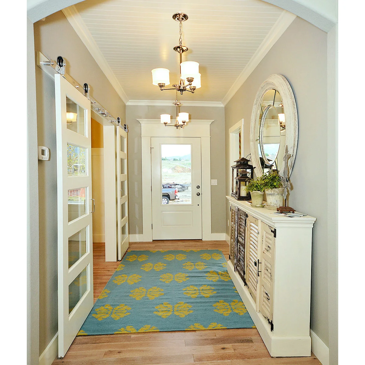 Narrow pale entrance hall with a glass-paneled front door, a shallow white cabinet, round mirror and colorful rug.