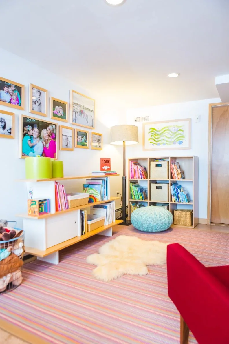 A white play corner has low wood-and-white book storage, a cube shelf with fabric bins, a pale blue pouffe, a striped rug and framed family photographs.