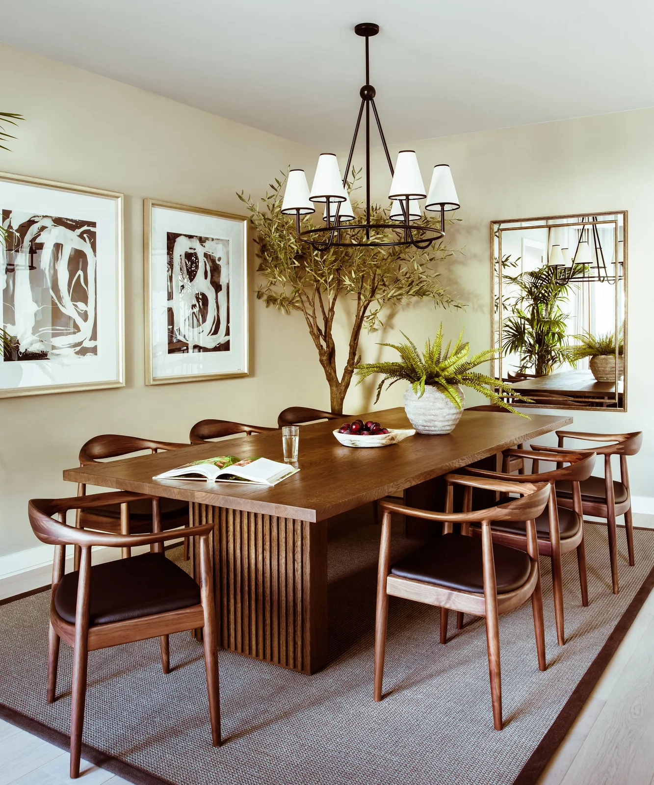 Curved wooden dining chairs surround a wooden table with a ribbed base beside framed art, a mirror and a shaded chandelier