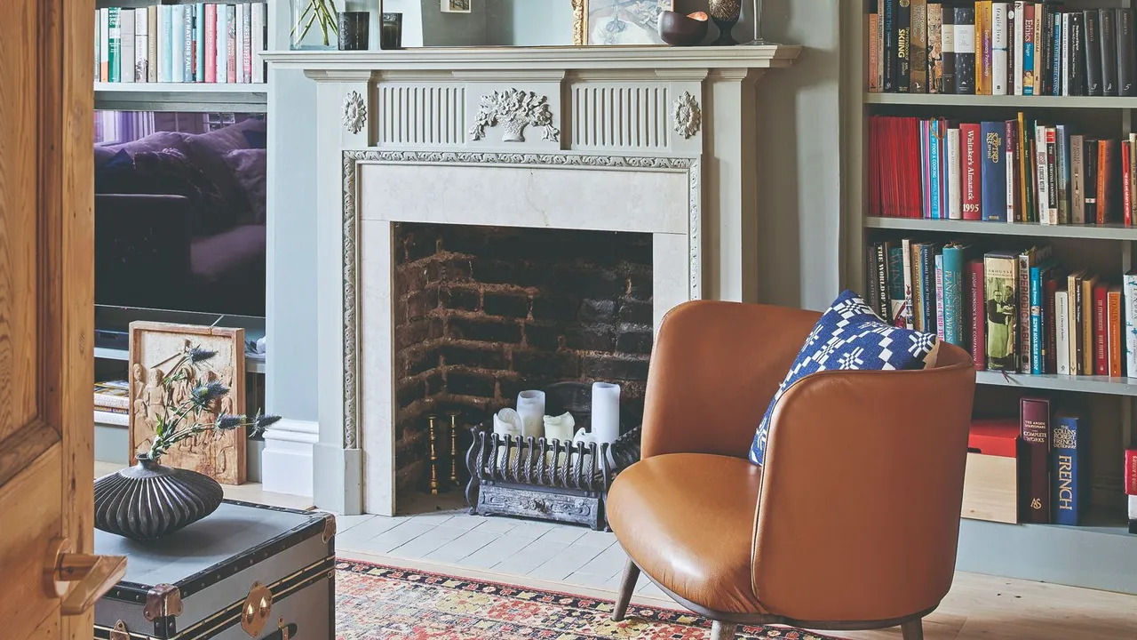 An ornate pale mantel sits between book-filled shelves beside a warm leather chair.