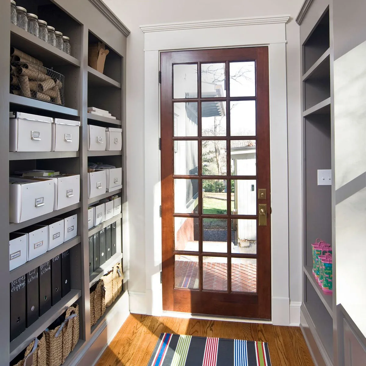 Narrow rear entrance with a glazed exterior door and fitted gray storage holding boxes, jars and rolled supplies.
