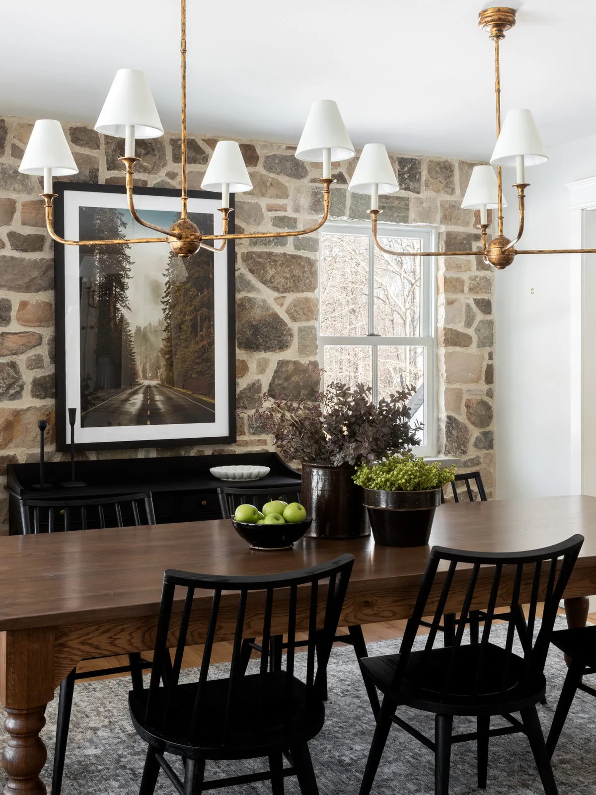 Dark chairs sit around a timber table beside a stone wall and warm chandelier.