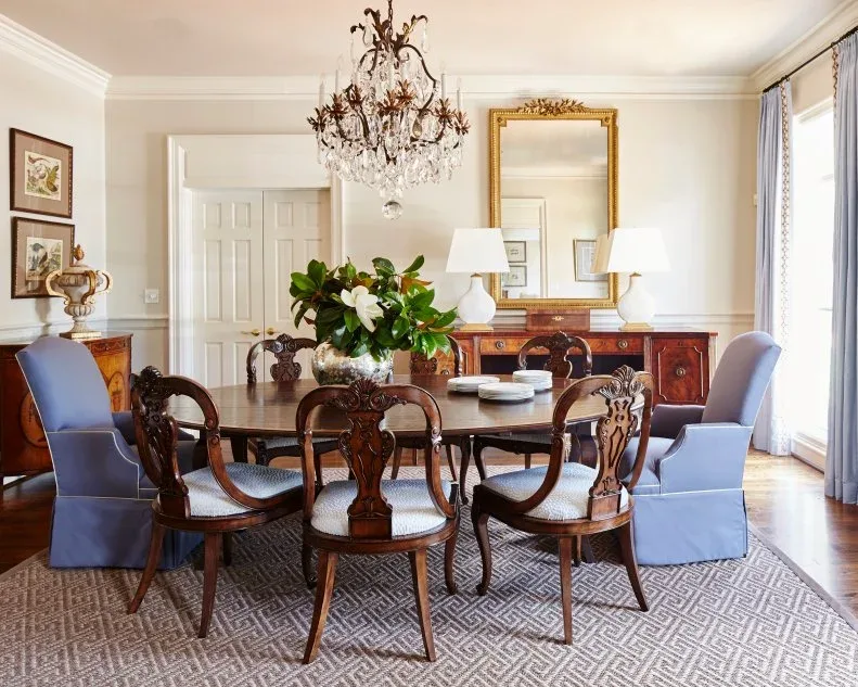 Large leafy arrangement with pale flowers on a dark dining table surrounded by carved chairs and blue end seats