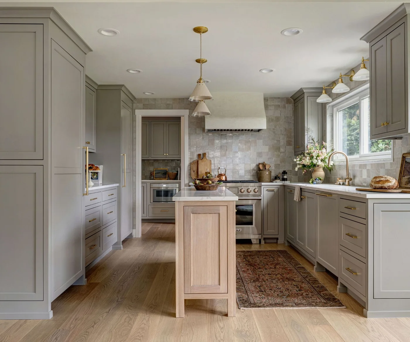 Warm gray paneled cabinets surround a narrow wooden island beside pale square tiles, gold-toned handles and a patterned runner