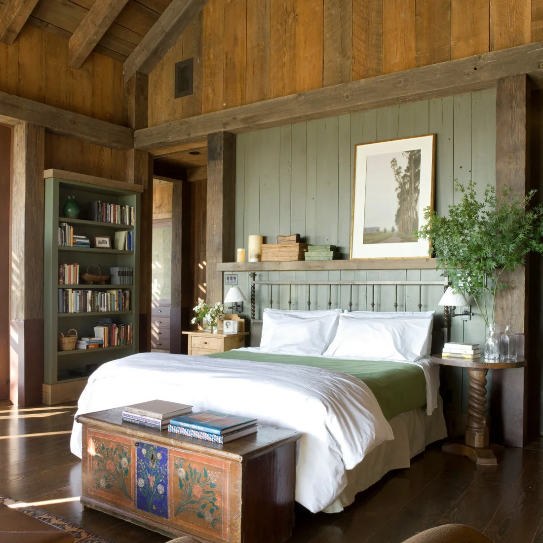 A green paneled bed wall with white bedding, dark metal headboard and painted chest beneath timber beams