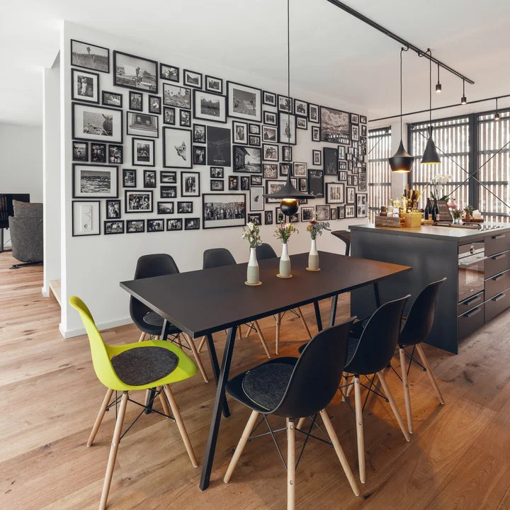 A dark rectangular table and mostly black chairs stand beneath a dense black-framed picture wall beside a kitchen island.