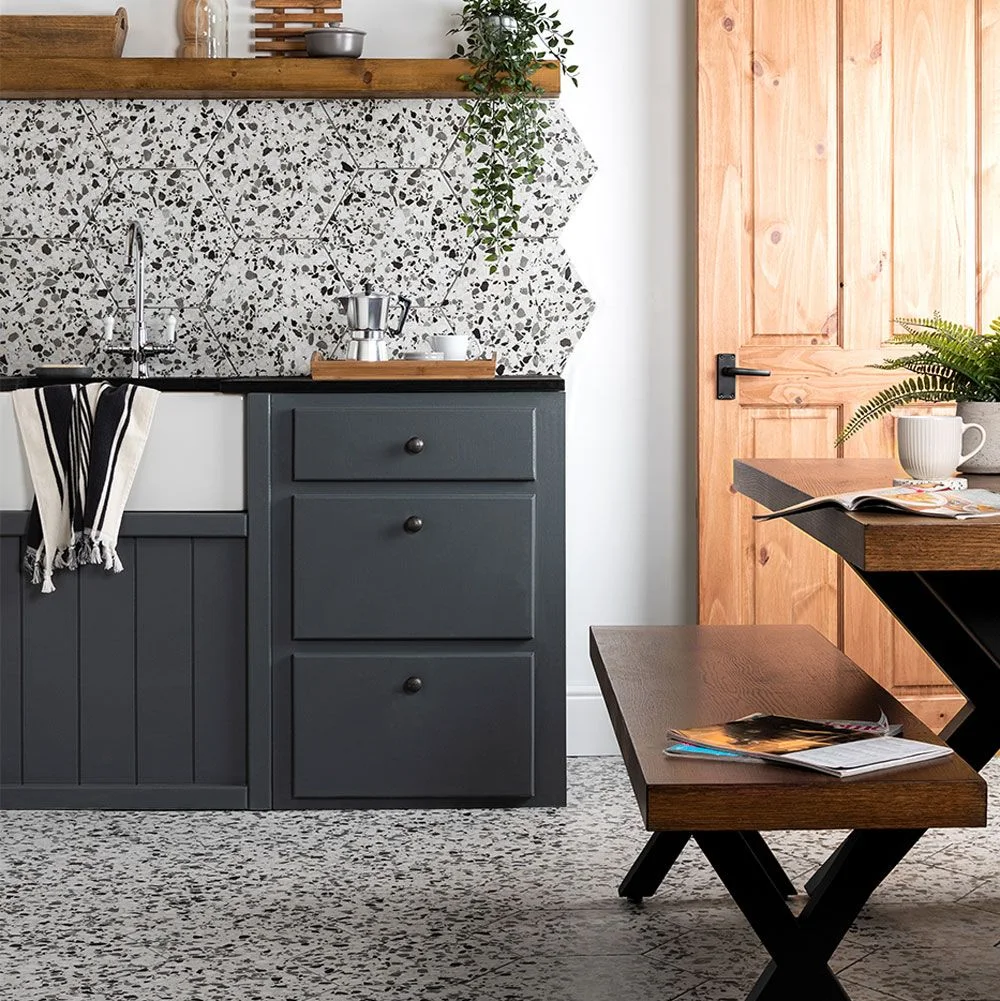 Charcoal gray drawers sit below a speckled geometric backsplash beside a white sink, wooden shelf and wooden door