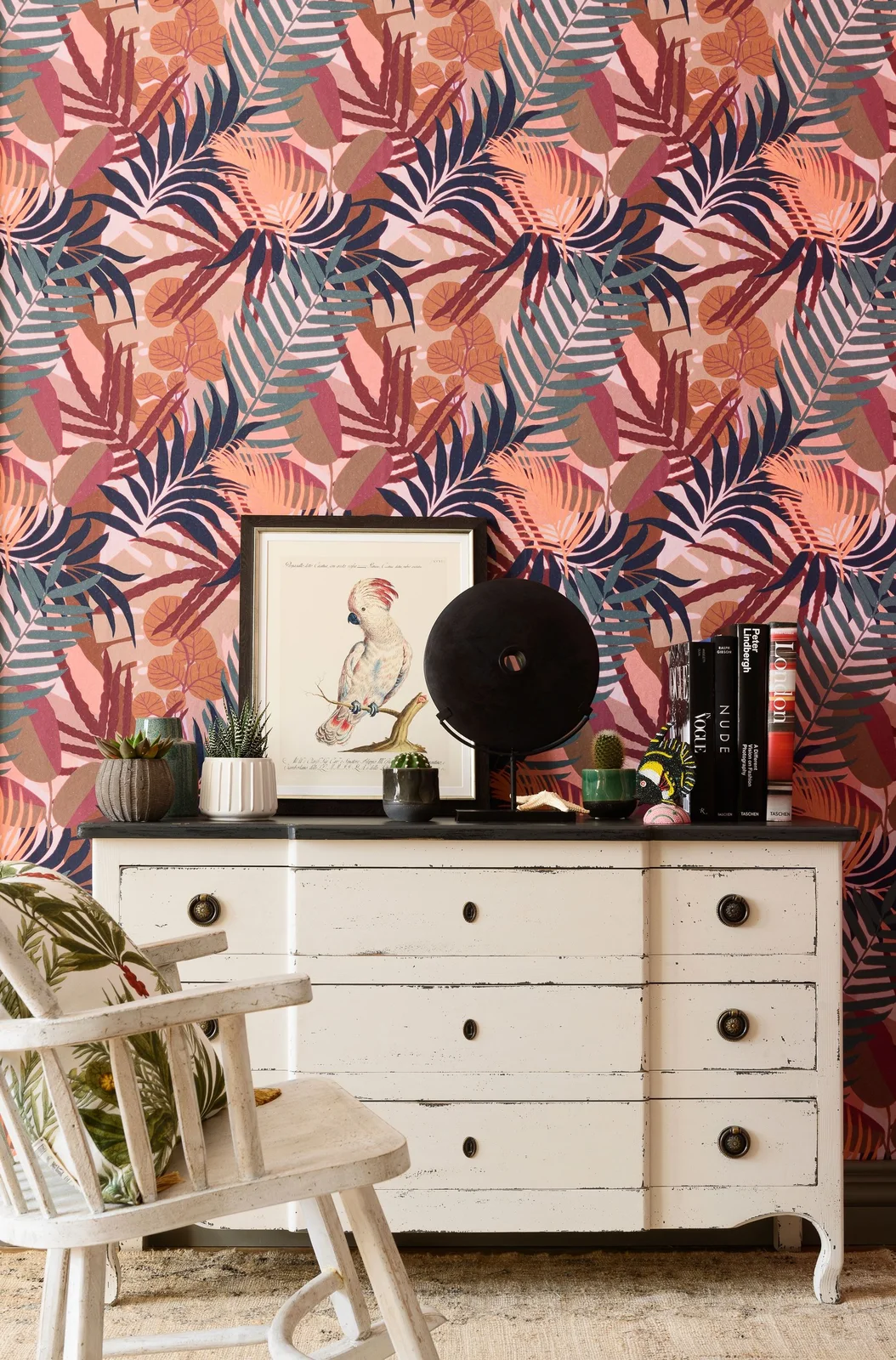 Pink and orange wallpaper with dark and teal palm leaves and pineapple motifs rises behind a white drawer chest, black fan and small white chair