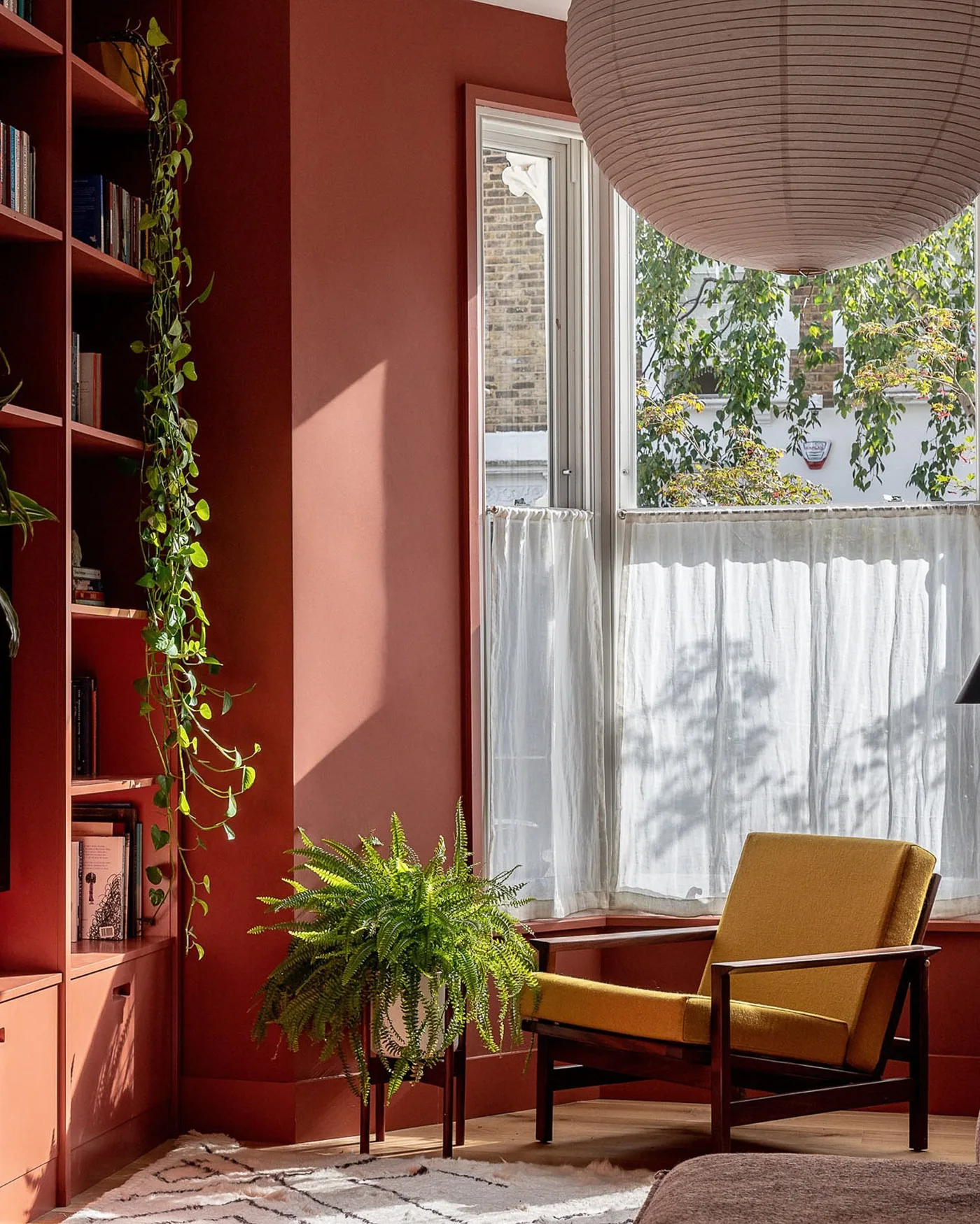 Warm red living room corner with matching bookcase, ochre armchair, pale window curtain and plants