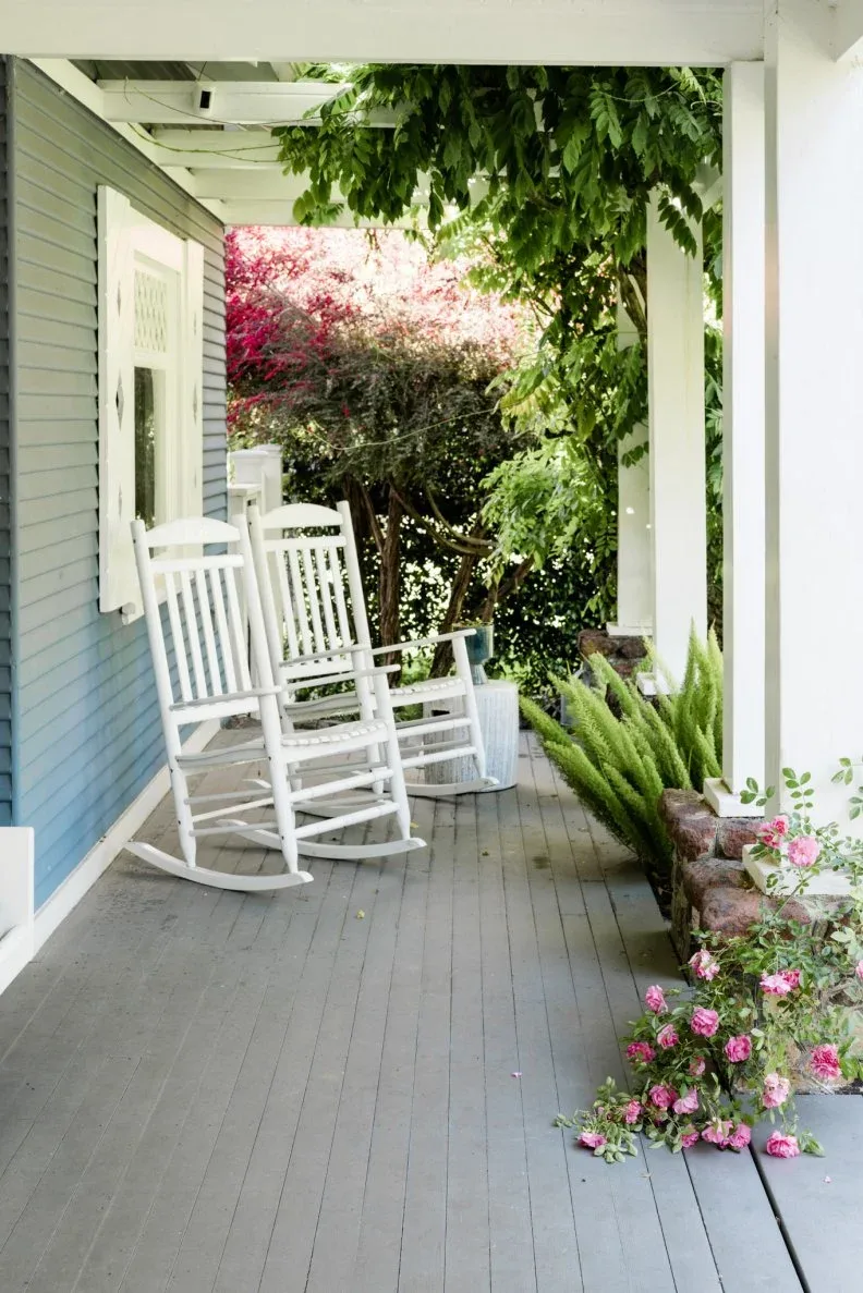 Two white rocking chairs along gray porch siding with greenery beside the outer columns
