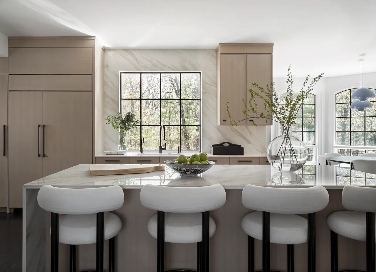 Beige cabinets face pale stools beside a black-framed window and dark worktop accents