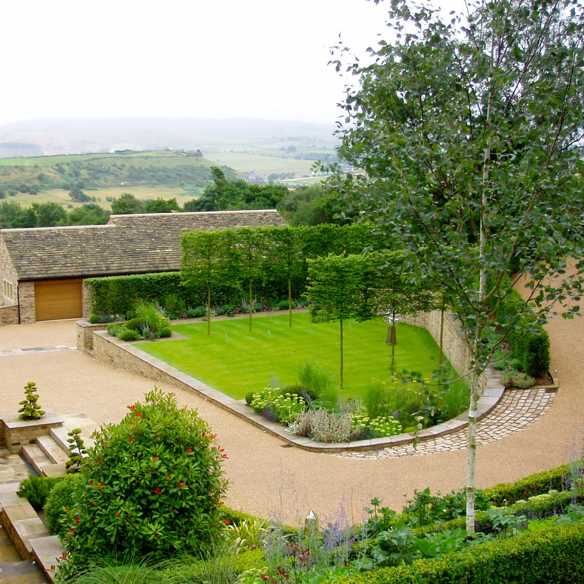 Pale driveway wrapping around a raised lawn island with small trees, planting beds and a low stone retaining wall.