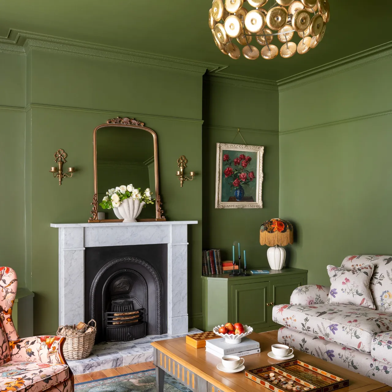 Olive green walls and ceiling surround floral seating, a wooden coffee table and a pale fireplace