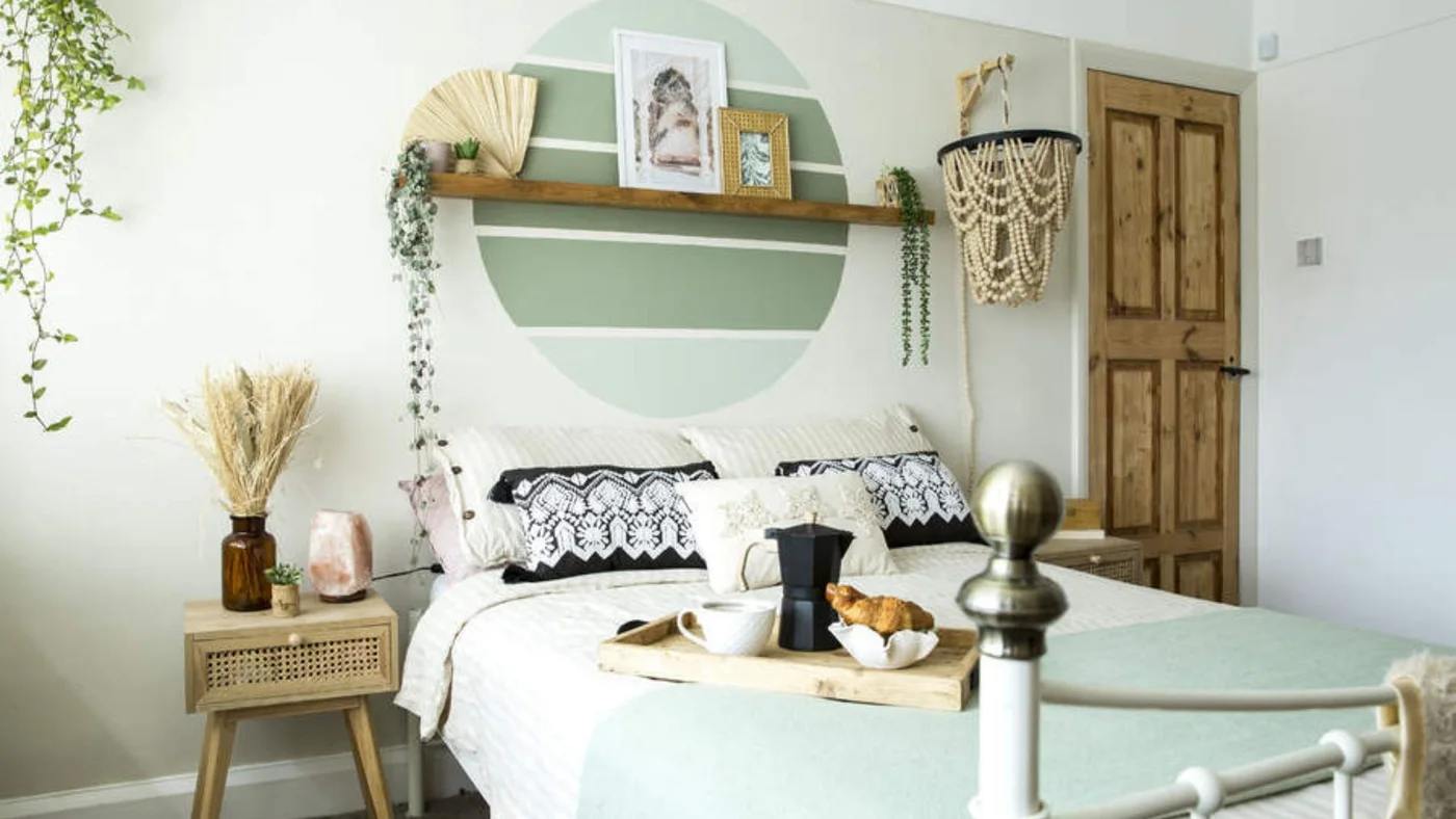 A striped green circle sits behind a wooden shelf above white bedding, patterned cushions and a pale metal bed frame