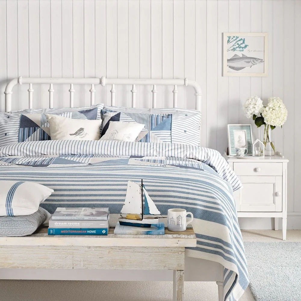 White paneled bedroom with a white metal bed, blue striped bedding, small nautical pictures and a pale wooden bench.