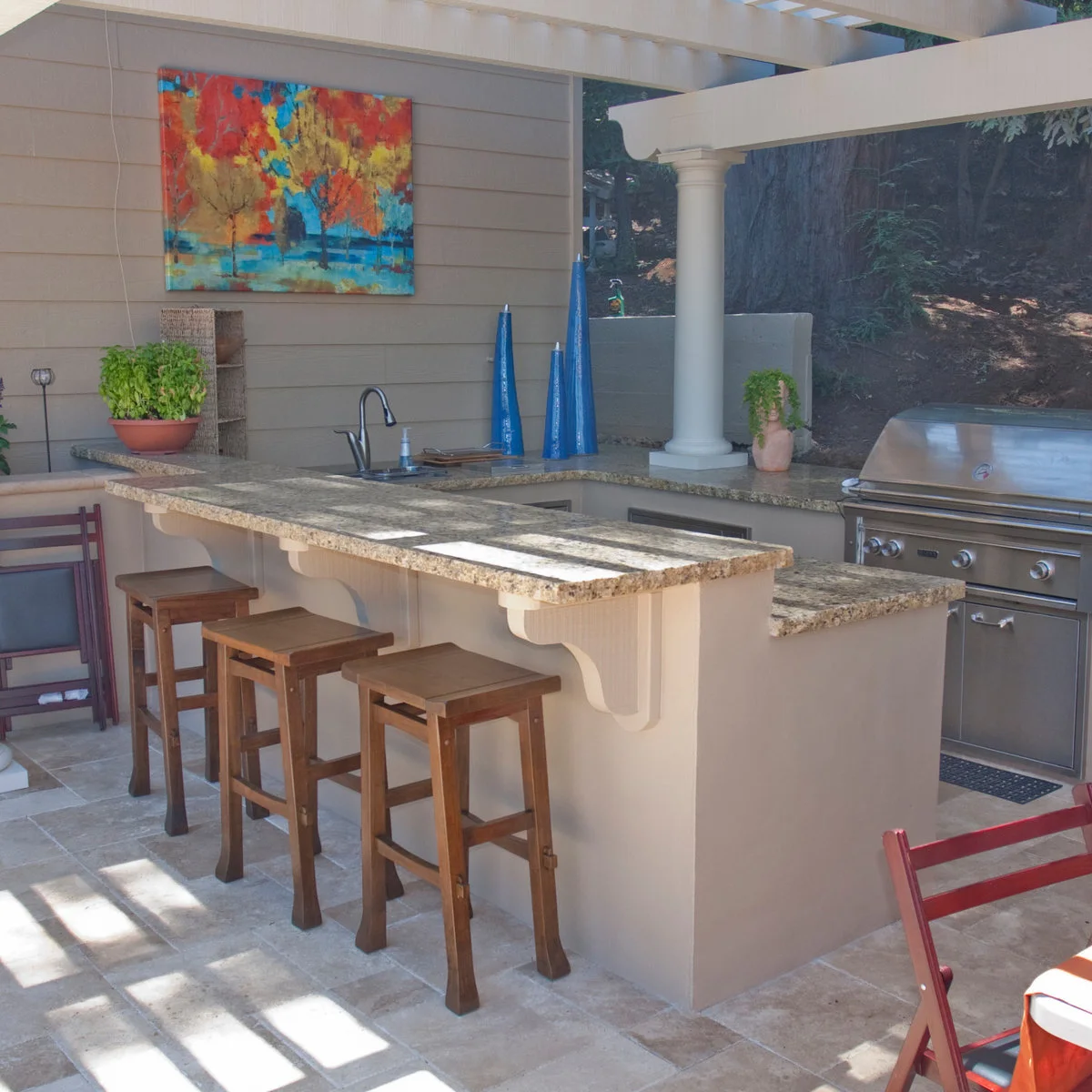 Outdoor kitchen with raised speckled counter ledge, three wood stools, sink, grill and colorful wall art