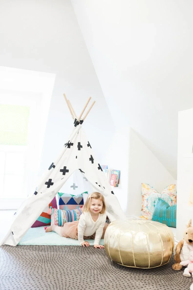 A child sits inside a white play tent patterned with black crosses, with colorful cushions, a pale blue floor pad and a round gold-colored pouffe beside it.