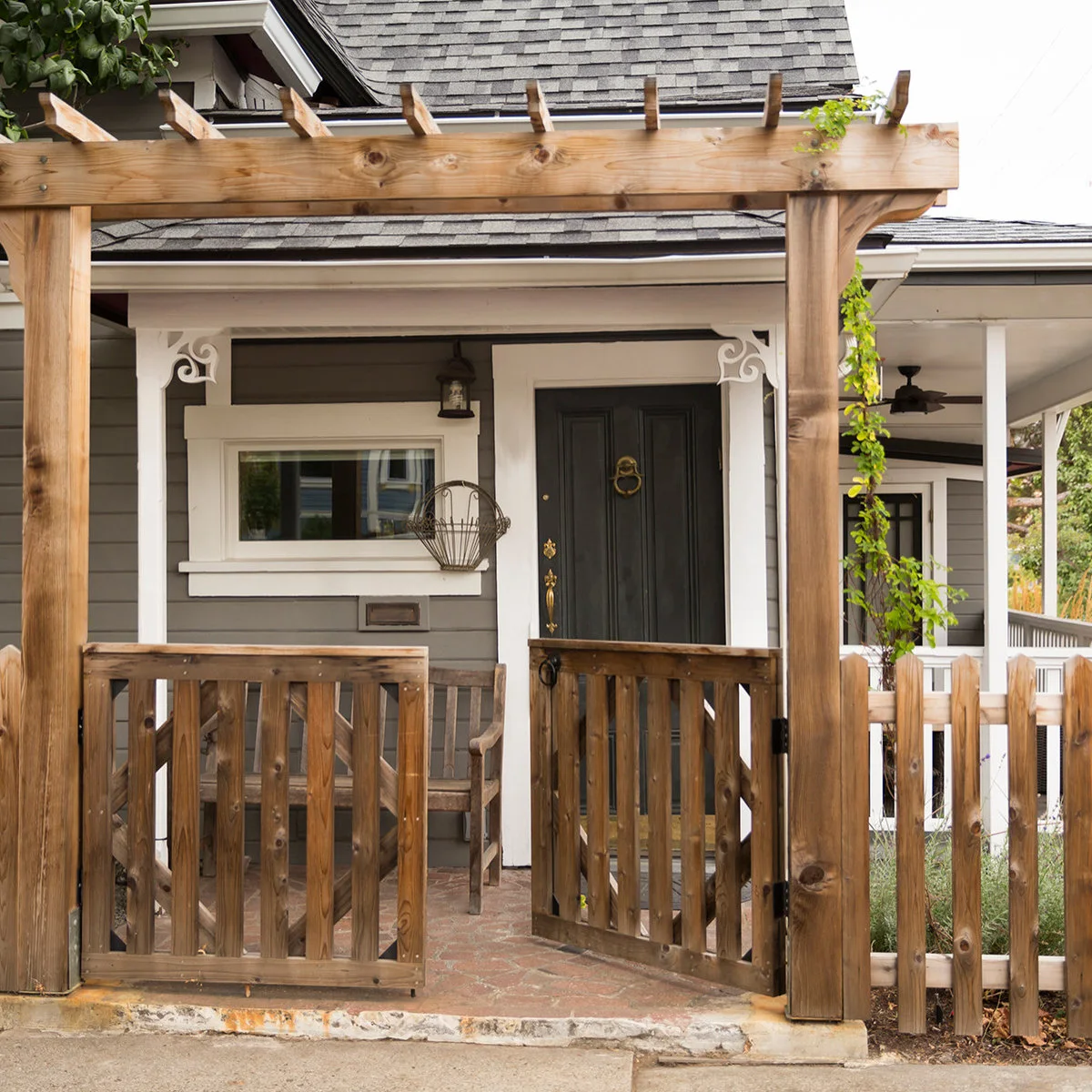 An open pair of wood gates beneath a simple timber arbor in front of a small house entrance.