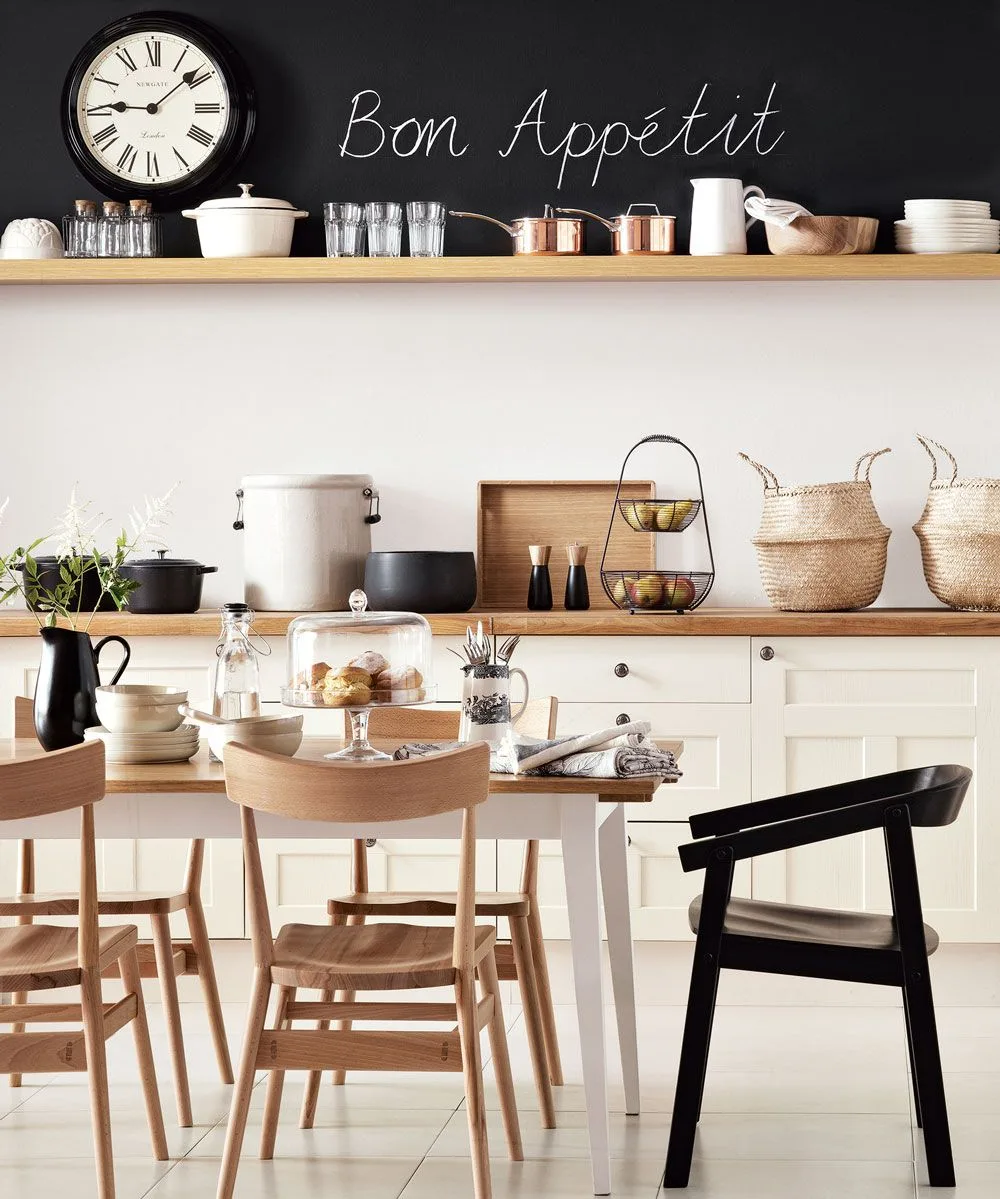 Cream framed base cupboards with round knobs sit below a timber shelf and dark chalkboard wall beside a dining table.